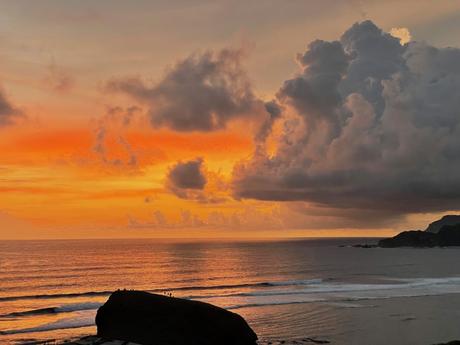 A vibrant orange sunset over the ocean with large clouds forming dramatic shapes in the sky, with small waves crashing onto the shore.