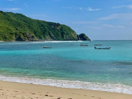 A turquoise bay with small wooden boats anchored near the shore. The steep, green cliffs rise dramatically from the water’s edge.