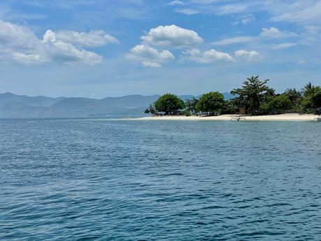 A peaceful coastal scene with calm, clear waters gently lapping a sandy beach. Distant mountains are faintly visible, creating a serene horizon.