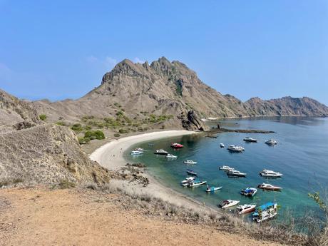 A scenic view of a secluded beach surrounded by rocky hills on Padar Island, Indonesia, with multiple boats anchored in the calm bay.