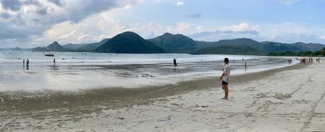 A wide sandy beach at low tide, with people walking along the shore and hills in the distance under a partly cloudy sky.