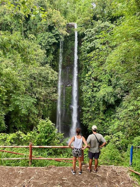 A couple stands in front of a lush, twin waterfall in a dense green jungle, holding hands and admiring the view.