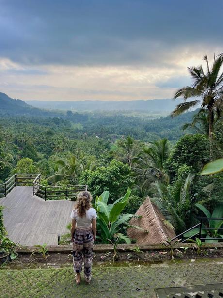 A woman standing on a deck overlooking a vast landscape of tropical forests and hills under a cloudy sky.