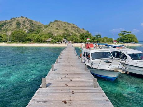 A wooden pier extending into crystal-clear turquoise waters with speed boats docked alongside, leading to a small island with hills in the background.