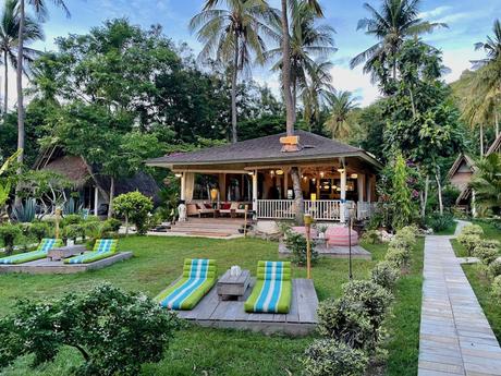 A tropical boutique hotel building with wooden lounge chairs on the front lawn, surrounded by tall palm trees and lush greenery. The home features a welcoming porch and well-kept garden.