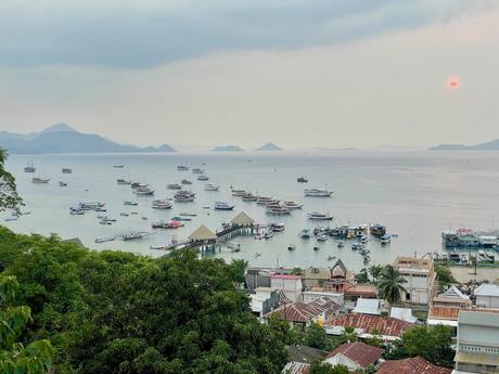 A view of a bay filled with boats in Labuan Bajo, Indonesia, with hills and islands in the background under an overcast sky.