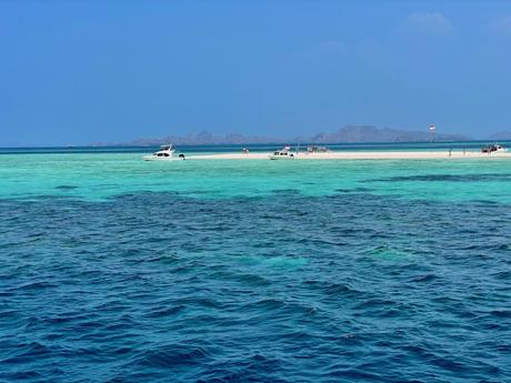 A small white boat docked on a bright turquoise sandbar in the middle of the ocean, surrounded by clear blue waters and distant mountains.