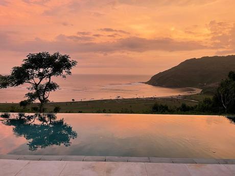 A breathtaking view of the sun setting over an infinity pool, with reflections of the orange and pink sky mirrored in the water, and the coastline and ocean in the distance.