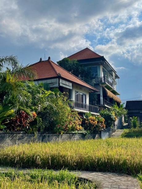 A traditional Balinese house with a red-tiled roof, surrounded by tropical plants and overlooking a green rice field.