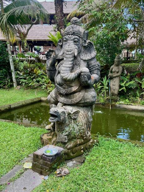 A stone statue of Ganesha, the Hindu elephant god, in a garden next to a pond, with lush plants and another small statue in the background.