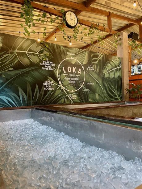 An ice bath filled with large ice cubes at a wellness center, with a mural on the wall behind it outlining steps for relaxation and recovery at LOKA Lounge.