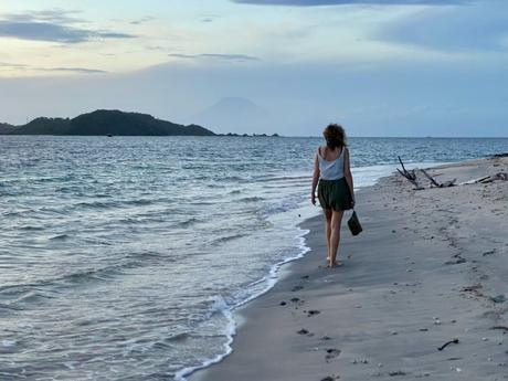 A woman walks barefoot along the shoreline at dusk, the ocean gently washing onto the beach. Distant islands and a silhouette of a mountain add depth to the tranquil scene.