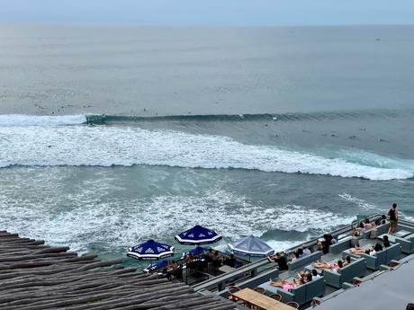 An elevated beachside café with people lounging and watching surfers riding waves in the ocean.