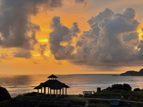 A silhouetted pavilion stands in front of a glowing orange sunset by the beach, with large, dark clouds towering above the ocean in the background.