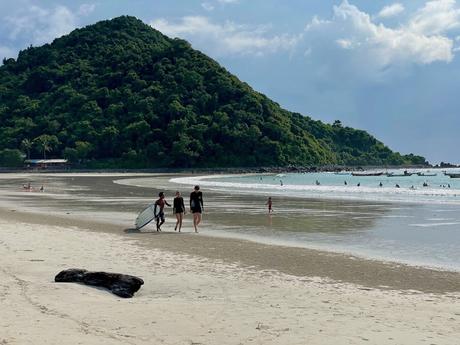 Three people, one holding a surfboard, walk along a tranquil beach with green, tree-covered hills in the background. The shoreline is dotted with small boats, and beachgoers are visible in the water.