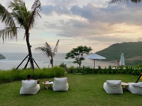 A relaxing outdoor seating area with white lounge chairs, overlooking a grassy lawn and a palm tree-lined coastline during sunset, with gentle ocean waves in the background.