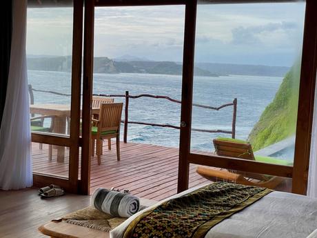 View from inside a room looking out onto a wooden deck with a table and chairs, overlooking the ocean and cliffs in the distance.