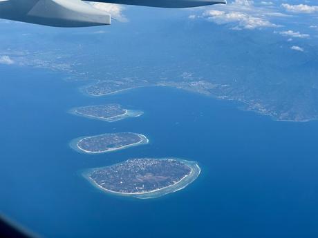 Aerial view from an airplane window showing a chain of small tropical islands (the Gili Islands) surrounded by turquoise waters, with the airplane wing visible in the top left corner.