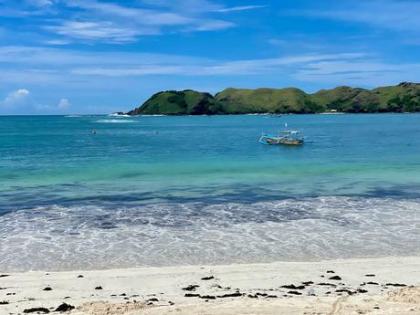 A small fishing boat anchored in crystal-clear turquoise water, with grassy hills in the background under a clear blue sky.