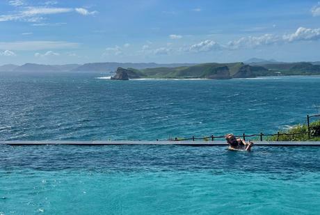 Alex Tiffany relaxing in an infinity pool with an expansive view of the ocean and distant cliffs under a bright blue sky.