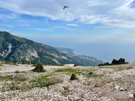 A view from a rocky hilltop looking out over a coastline. A paraglider soars through the sky above the distant beach and ocean, with mountains in the background.