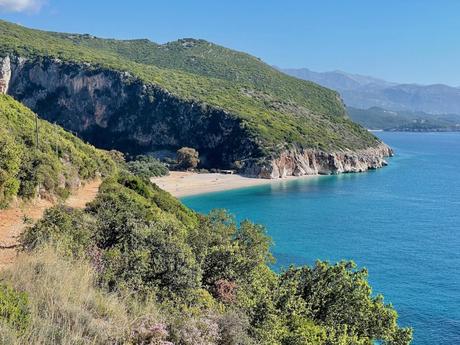 A scenic view of a secluded beach with cliffs and dense greenery on one side. The clear blue waters stretch to the horizon, with a few small figures seen enjoying the sandy shore.