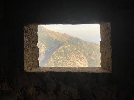 A view through a small rectangular window in an Albanian bunker, showing a mountainous landscape with rugged peaks and green forests.