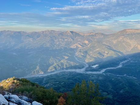 A mountain range in Albania bathed in soft, late-afternoon light. The valley below is crisscrossed by a winding river, with various shades of blue and green in the rolling landscape.