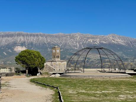 A large open area with a stone clock tower and the framework of an amphitheater. Behind, rugged mountains rise under a bright blue sky.