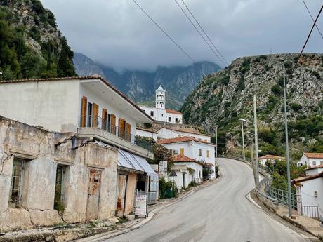 The small village of Dhermi nestled against rocky hills, with rustic houses and a white church tower visible under a cloudy sky.