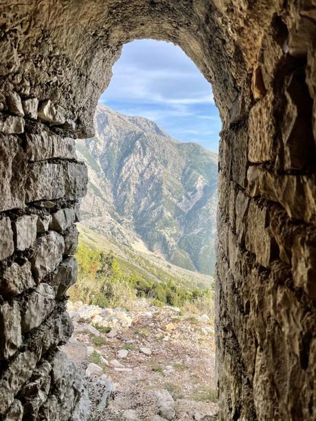 Looking out from within a stone tunnel, framed by rough rocks, revealing a view of a steep, mountainous terrain under a bright, clear sky.