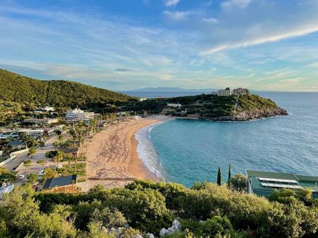 A scenic view of a crescent-shaped beach with golden sand and turquoise waters, surrounded by green hills and a small seaside village.