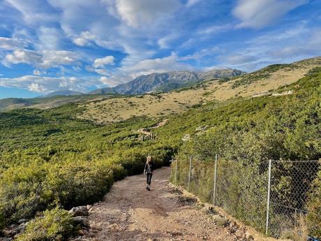 A person walking along a dirt path bordered by greenery, with rolling hills and mountains in the distance under a partly cloudy sky.