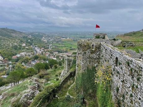 An ancient stone fortress with a red Albanian flag flying above, overlooking a valley with green hills and scattered villages.