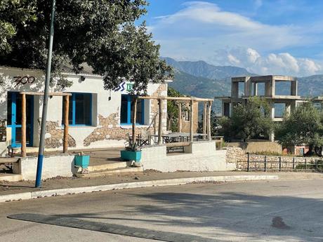 A rural building in Himare, Albania with blue accents stands near the road, with an unfinished concrete building behind it and a backdrop of mountains.