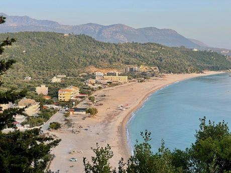 A view of a coastal town nestled between the mountains and the sea, with a long sandy beach curving along the shoreline. The town is surrounded by greenery and hills.