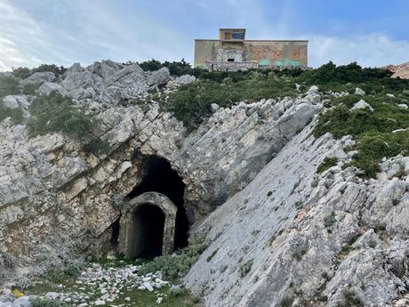 An entrance to a bunker tunnel built into rocky terrain. Above the cave, a small building is perched with graffiti on its walls.