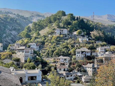 The hillside city of Gjirokaster nestled among lush greenery, with traditional stone houses scattered across the slope, surrounded by trees and mountains in the background.