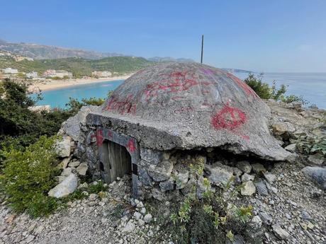 A small concrete bunker with graffiti stands on a hillside overlooking a turquoise blue bay, with mountains in the distance and a beachside town below.