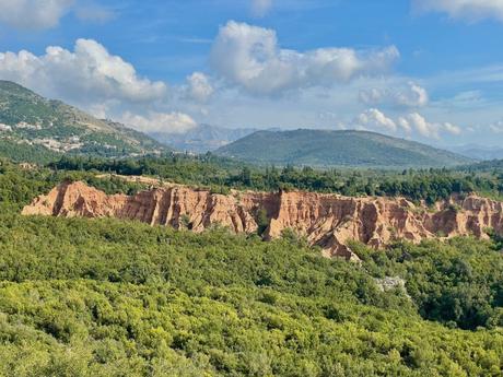 A dramatic landscape with jagged red rock formations standing out against a lush green valley. The sky above is dotted with white clouds.