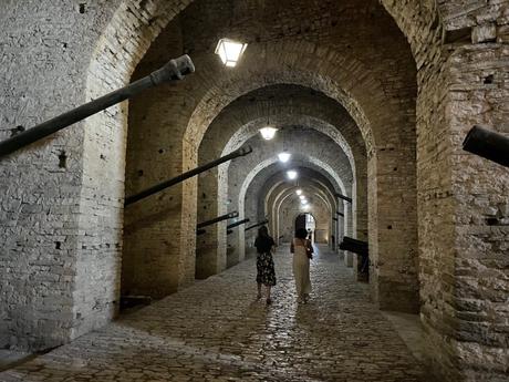 Two people walking through a long, arched stone tunnel lit by lanterns inside Gjirokaster castle. The stonework has a medieval feel, with cannons lining the walls.