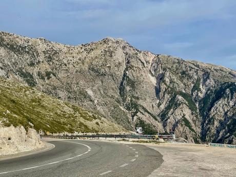 A road winding through a mountainous landscape on the Albanian Riviera, with steep, rocky slopes on either side. The mountains appear barren, with minimal vegetation.