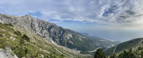 A panoramic shot of a mountain range with jagged cliffs and a view of a winding road along the coastline below. The sky is cloudy, with patches of sunlight on the landscape.