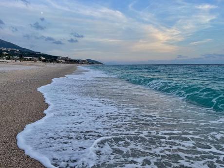 The shoreline of a beach at dusk, with gentle waves lapping against the pebbly shore. The sky is tinged with soft pastel colors as the sun sets.