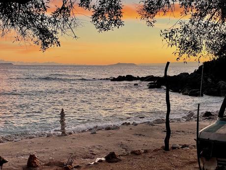 A peaceful beach at sunset, with a stack of balanced stones near the water’s edge, framed by overhanging tree branches and vibrant orange and pink hues in the sky.