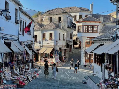 A cobbled street in the Old Bazaar of Gjirokaster lined with small shops selling traditional crafts and textiles. People are seen walking and exploring the market, with vibrant red and white buildings in view.