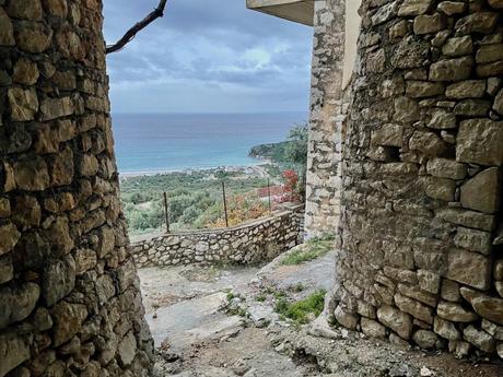 A view from between two old stone walls, looking out over a beach and the ocean in the distance, framed by greenery and some red flowers.