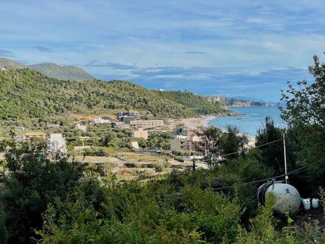 An Albanian coastal village nestled between green hills and the shoreline. Houses and buildings are visible, along with a curving coastline stretching into the distance.