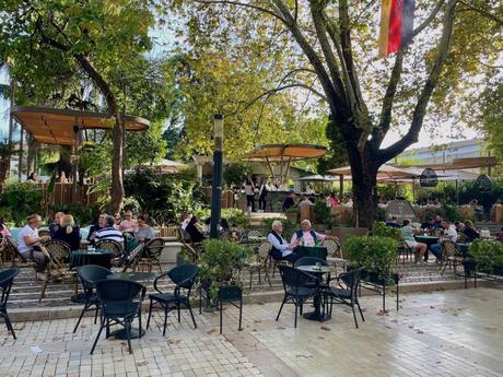 A lively outdoor cafe scene with people seated under large trees, enjoying their time in a shaded garden area. The setting feels relaxed, with greenery and outdoor furniture adding to the atmosphere.