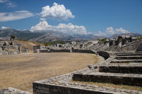 A wide view of the ancient amphitheater ruins at Salona, Croatia, with stone walls and steps under a bright blue sky, framed by mountains in the distance.